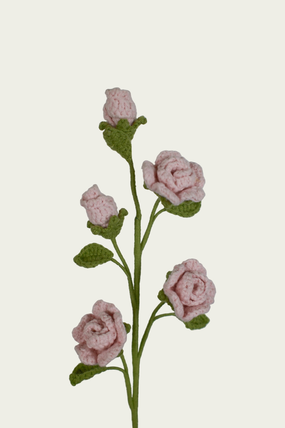 A crocheted artificial flower stem featuring five light pink roses with green leaves and buds, set against a plain light gray background.